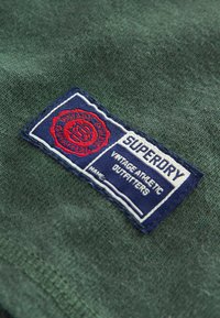 Green cotton fabric with a woven blue patch. The patch features the text “SUPERDRY” and “VINTAGE ATHLETIC OUTFITTERS” in white and red.