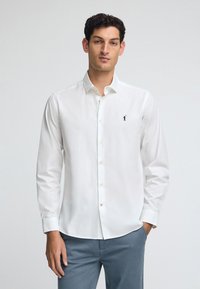 Vit skjorta med knappar gjord av bomull; har långa ärmar, en spetsig krage och en liten marinblå logga på vänster bröst. Lätt textur.