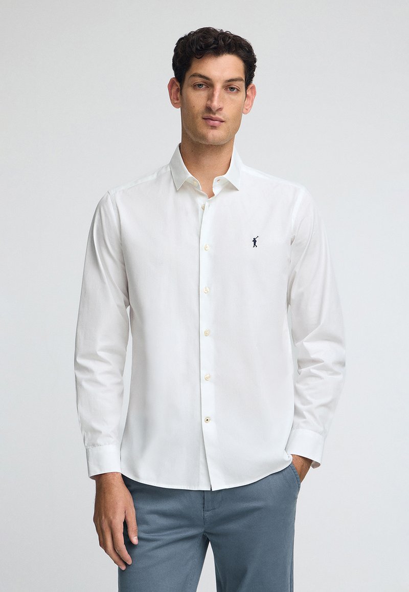 Vit skjorta med knappar gjord av bomull; har långa ärmar, en spetsig krage och en liten marinblå logga på vänster bröst. Lätt textur.