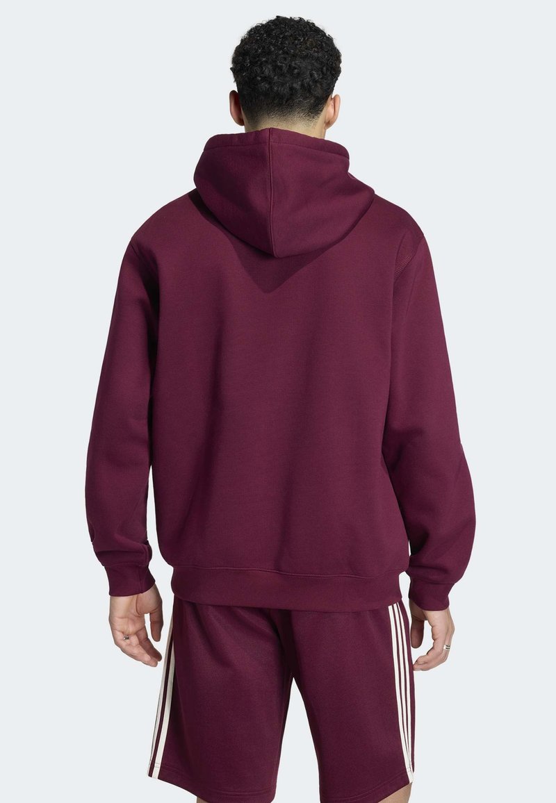 Uomo che indossa una felpa con cappuccio color amaranto e pantaloncini abbinati con strisce bianche laterali, in piedi con la schiena rivolta verso l'osservatore.