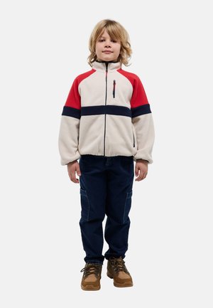 Enfant debout portant une veste polaire crème avec des rayures rouges et noires, un pantalon cargo foncé et des chaussures de randonnée marron sur un fond blanc.