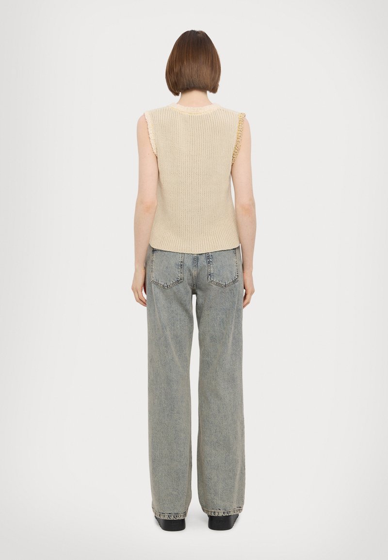 Beige gebreid mouwloos topje met een geribbelde textuur, voorzien van gerimpelde randen, gecombineerd met lichtblauwe wijde jeans en donkere schoenen.