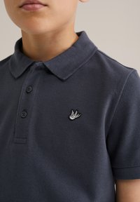 Námornícky polo tričko vyrobené z textúrovaného materiálu s golierom, tromi gombíkmi a bielym vyšitým logom vtáka na hrudi.