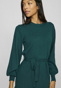 Femeie îmbrăcată într-o rochie tricotată cu manșete lungi, de culoare verde închis, cu mâneci tip balon și un cordon legat la talie, privind în lateral.