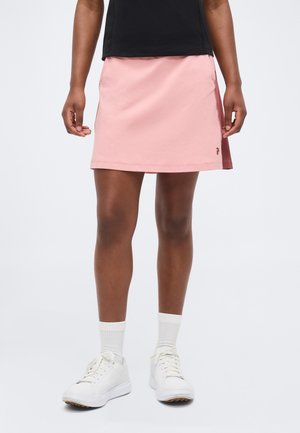 Άτομο που φοράει μαύρο τοπ, ανοιχτό ροζ φούστα, λευκές κάλτσες και λευκά αθλητικά παπούτσια, στεκόμενο μπροστά σε απλό ανοιχτό φόντο.