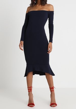 Vrouw draagt een marineblauwe midi-jurk met lange mouwen en een off-shoulder ontwerp, met een gerimpelde zoom en rode hoge hakken met veters.