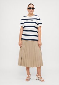 Stribet navy og hvid t-shirt med "Tommy Hilfiger" tekst, parret med en plisseret beige nederdel. Fødderne i hvide strop sandaler med hæl.