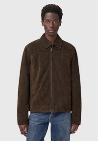 Mand iført en mørkebrun ruskindsjakke med lynlås og krave samt blå jeans, stående mod en ensfarvet lys grå baggrund.