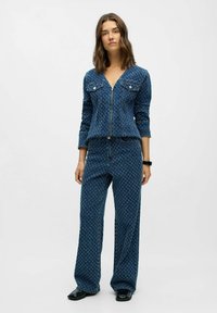 Mujer vestida con una chaqueta de mezclilla azul con estampado y cremallera, acompañada de unos pantalones anchos a juego, de pie frente a un fondo blanco.