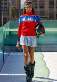 Maglione a collo alto rosso e blu con la scritta "Aptes SKI", gonna bianca plissettata con motivo a griglia, stivali da neve neri e lucidi, e un casco nero.