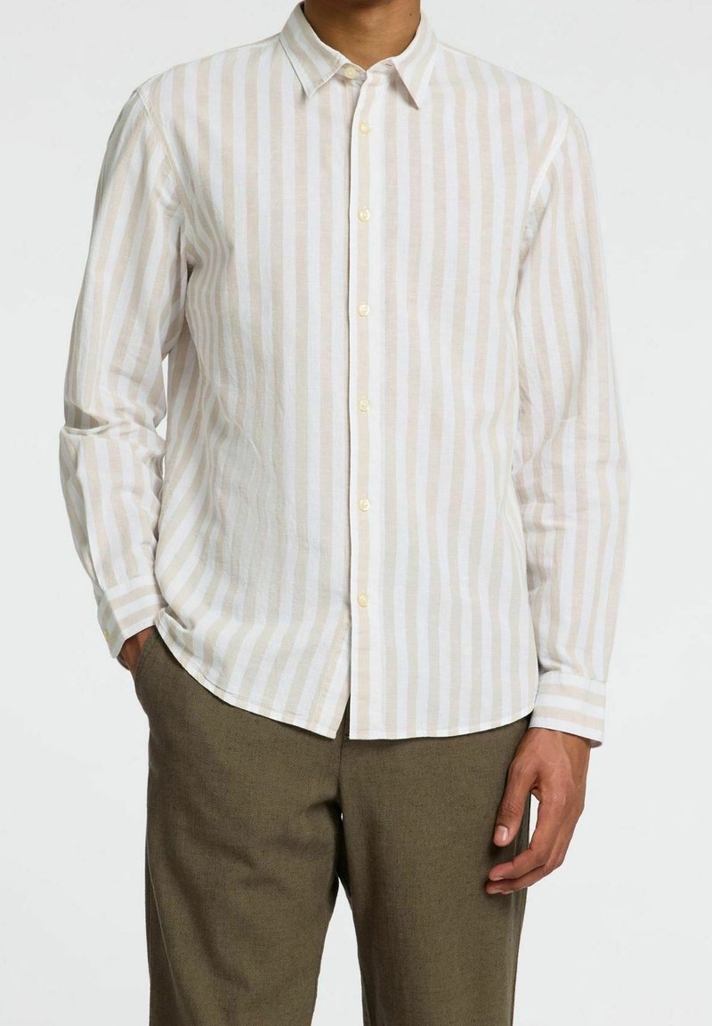 Homme portant une chemise beige et blanche à rayures verticales boutonnée avec manches longues et un pantalon marron, debout avec une main dans la poche.