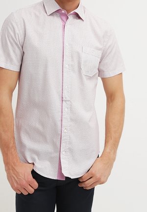 Homme portant une chemise blanche à manches courtes avec un petit motif géométrique rose, poche poitrine gauche, et pantalon noir, debout, mains près des poches.