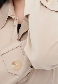 Beige trenchcoat med bred krage, knappstängning och detaljer i guldton. Mjuk tyg med synliga söms detaljer.
