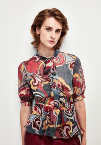 Blusa peplum multicolore con motivi floreali in tonalità di rosso, giallo e blu, dotata di una chiusura con bottoni e dettaglio con nodo al collo.