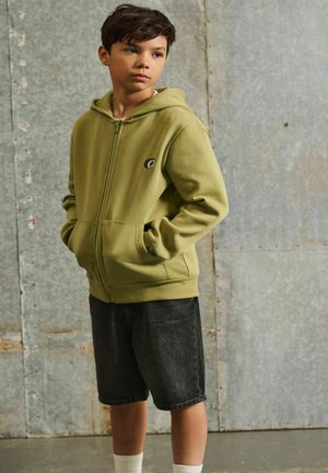 Fant fant z olivno zeleno jopico na zadrgo in črnimi jeans kratkimi hlačami, stoji z rokami v žepih ob sivi industrijski steni.