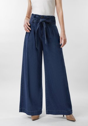 Žena má oblečené tmavomodré široké nohavice s vysokým pásom a viazaným opaskom, v kombinácii s béžovými topánkami so špicatou špičkou a bielym tielkom bez rukávov.