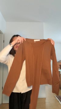 Persona che tiene in mano un cardigan marrone a maniche lunghe in una stanza ben illuminata con pareti bianche e pavimento in legno.