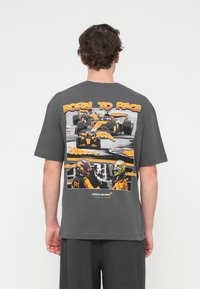 Γκρι βαμβακερό μπλουζάκι με μεγάλο γραφικό τύπωμα που απεικονίζει αυτοκίνητα και οδηγούς της Formula 1. Κείμενο "BORN TO RACE" τονισμένο με πορτοκαλί χρώμα. Κοντά μανίκια.