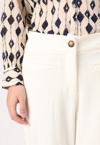 Vita texturerade byxor med en synlig knapp, matchade med en mönstrad blus med marinblå och beige diamantformer.