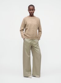 Γυναίκα στέκεται με τα χέρια στις τσέπες φορώντας μπεζ πουλόβερ, φαρδιά ανοιχτόχρωμα χακί παντελόνια και ανοιχτόχρωμα μυτερά παπούτσια σε λευκό φόντο.