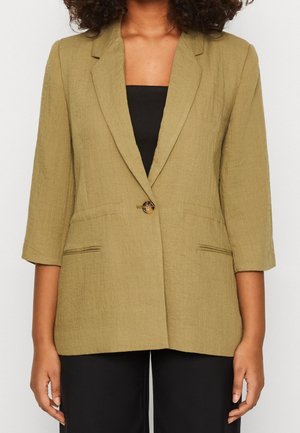 Vrouw die een licht olijfgroene blazer draagt met driekwart mouwen, een enkele schildpadknop, over een zwart top en zwarte broek.