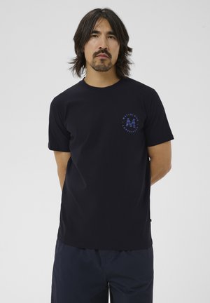 Muž so stredne dlhými vlasmi, ktorý má na sebe tmavomodré tričko s malým logom Matinique Copenhagen na ľavej strane hrudníka, stojaci pred jednoduchým pozadím.