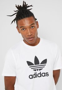 Fehér pamut póló, középen egy nagy fekete háromcsíkos Adidas logóval és levélmintával. Rövid ujjú, kerek nyakkivágású.
