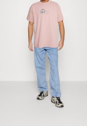 T-shirt en coton rose clair avec un petit imprimé graphique sur la poitrine, accompagné d'un jean en denim bleu à coupe ample et de baskets à motifs.