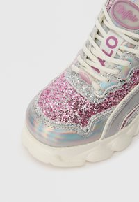Χοντρό sneaker με λευκή σόλα, ροζ και ασημένιο γκλίτερ επάνω μέρος, ιριδίζουσες πάνελ, κρεμ λουριά, και το λογότυπο "Buffalo" στη γλώ σσα και στην αγκράφα του λουριού.