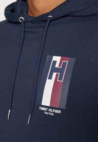 Sudadera de color azul marino con un logo frontal que presenta una gran 'H' en franjas de color blanco, rojo y azul. Incluye ojales de metal y cordones.