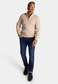 Beige ribbelgebreide pullover met een halve rits kraag. Gecombineerd met donkerblauwe jeans en donkere sneakers, wat een casual uitstraling geeft.