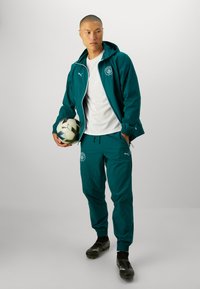 Completo tuta verde acqua con giacca con cappuccio e pantaloni, caratterizzato da polsini elastici, dettagli del logo bianco e una maglietta bianca sotto. Tiene in mano un pallone da calcio.