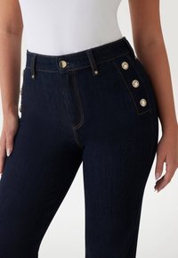 Dunkelblaue Denim-Jeans mit hoher Taille, ausgestattet mit goldenen Knöpfen an der Seite und kontrastierenden Nähten für zusätzlichen Detailreichtum und Textur.