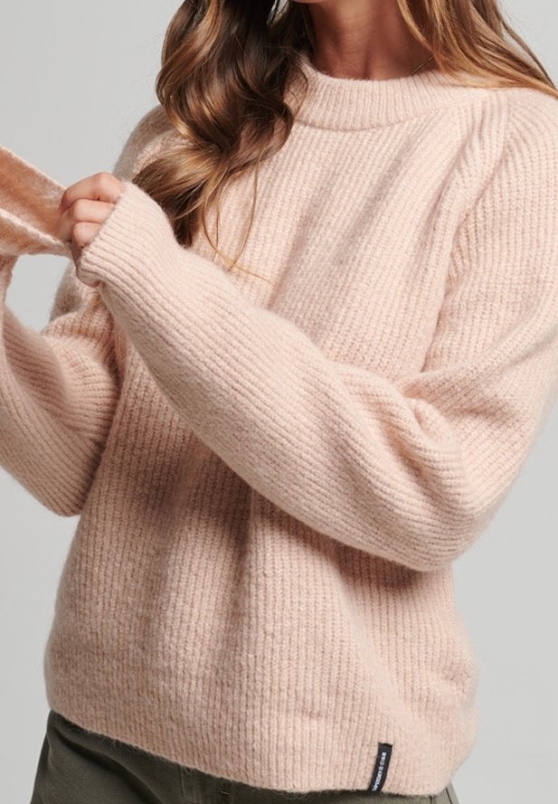 Femme portant un pull côtelé doux rose pâle, ajustant le poignet de la manche, avec de longs cheveux ondulés et un pantalon vert olive visible.