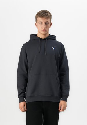 Young man wearing a black hoodie with a small blue and white bird logo on the chest, standing against a plain white background.