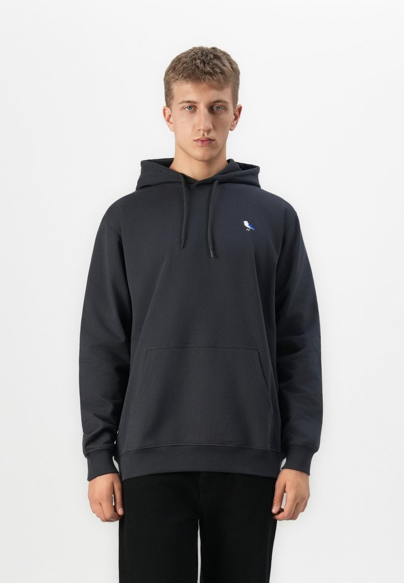 Young man wearing a black hoodie with a small blue and white bird logo on the chest, standing against a plain white background.