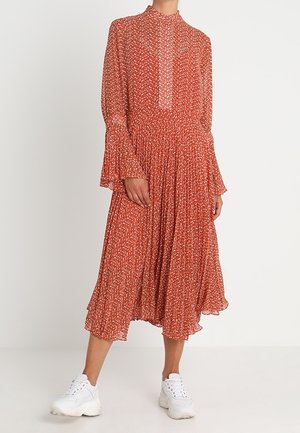 Femme portant une robe midi à manches longues couleur rouille avec un petit imprimé floral blanc et des baskets blanches sur un fond uni.