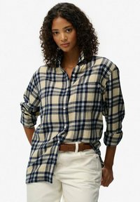 Chemise en flanelle à carreaux beiges et bleus marine, avec un design à boutons, des manches longues retroussées et une poche sur la poitrine. Portée sur un pantalon blanc.