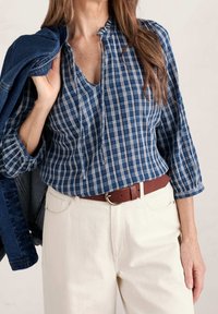 Blouse à carreaux bleus et blancs avec un col en trou de serrure, manches trois-quarts, portée avec un pantalon crème et une ceinture marron. Veste en denim portée sur l'épaule.