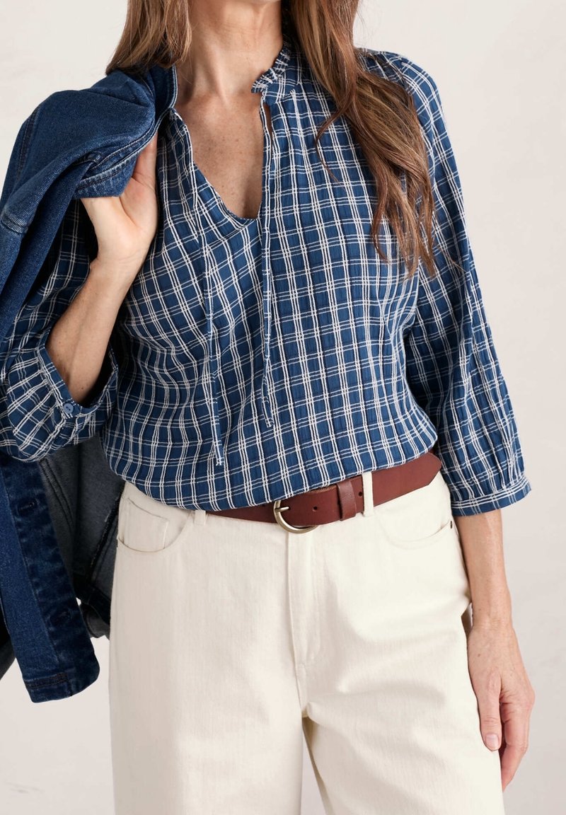 Blouse à carreaux bleus et blancs avec un col en trou de serrure, manches trois-quarts, portée avec un pantalon crème et une ceinture marron. Veste en denim portée sur l'épaule.