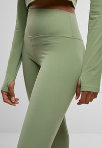 A close-up of light green leggings made of stretch fabric, featuring a smooth texture and high waistband. Long sleeves with thumbholes visible.