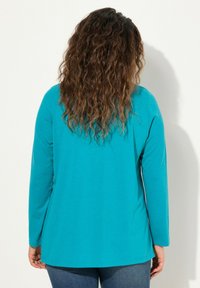 Teal lange mouw shirt met een ontspannen pasvorm, achteraanzicht dat een gladde textuur en eenvoudig ontwerp toont, gecombineerd met blauwe jeans.