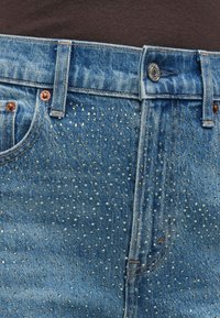 Τζιν παντελόνια σε ανοιχτό μπλε χρώμα με μεταλλικές λεπτομέρειες κουκκίδων διασκορπισμένες μπροστά, με εξαρτήματα σε χάλκινη απόχρωση και κούμπωμα με κουμπί.