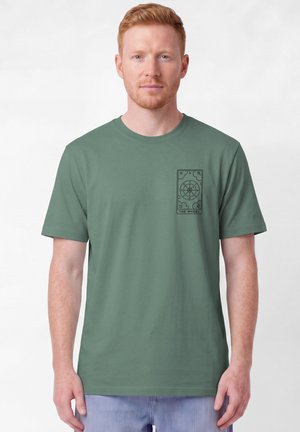 Rothaariger Mann, der ein schlichtes grünes T-Shirt mit schwarzem Tarotkartendesign mit der Aufschrift "The Wheel" auf der linken Brust trägt und vor weißem Hintergrund steht.