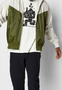 Giacca a vento verde e crema con chiusura con zip, polsini elastici e orlo a coste. La maglietta bianca sottostante presenta una stampa grafica.