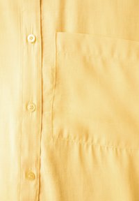 Camicia gialla con bottoni, dal tessuto liscio, con taschino sul petto e cinque bottoni. Il tessuto ha una leggera lucentezza.