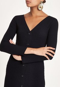 Robe en maille côtelée noire avec un décolleté en V, des manches longues et une patte de boutonnage sur le devant. Tissu texturé et coupe ajustée.