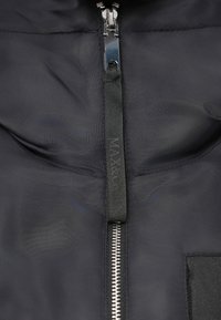 Jachetă din țesătură neagră, cu un fermoar frontal și un tab de prindere texturat, având o mică emblemă. Material neted, cu o strălucire subtilă.