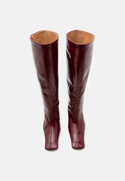 Λουστραριστές μπορντό δερμάτινες μπότες μέχρι το γόνατο με τετράγωνα δάχτυλα, όρθιες πάνω σε απλό λευκό φόντο.