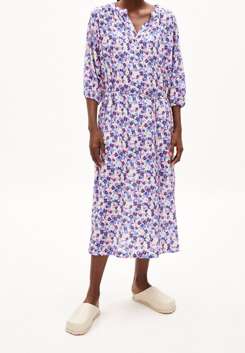 Personne portant une robe midi fleurie avec manches trois-quarts et un lien sur le côté, associée à des chaussures beiges claires à enfiler.